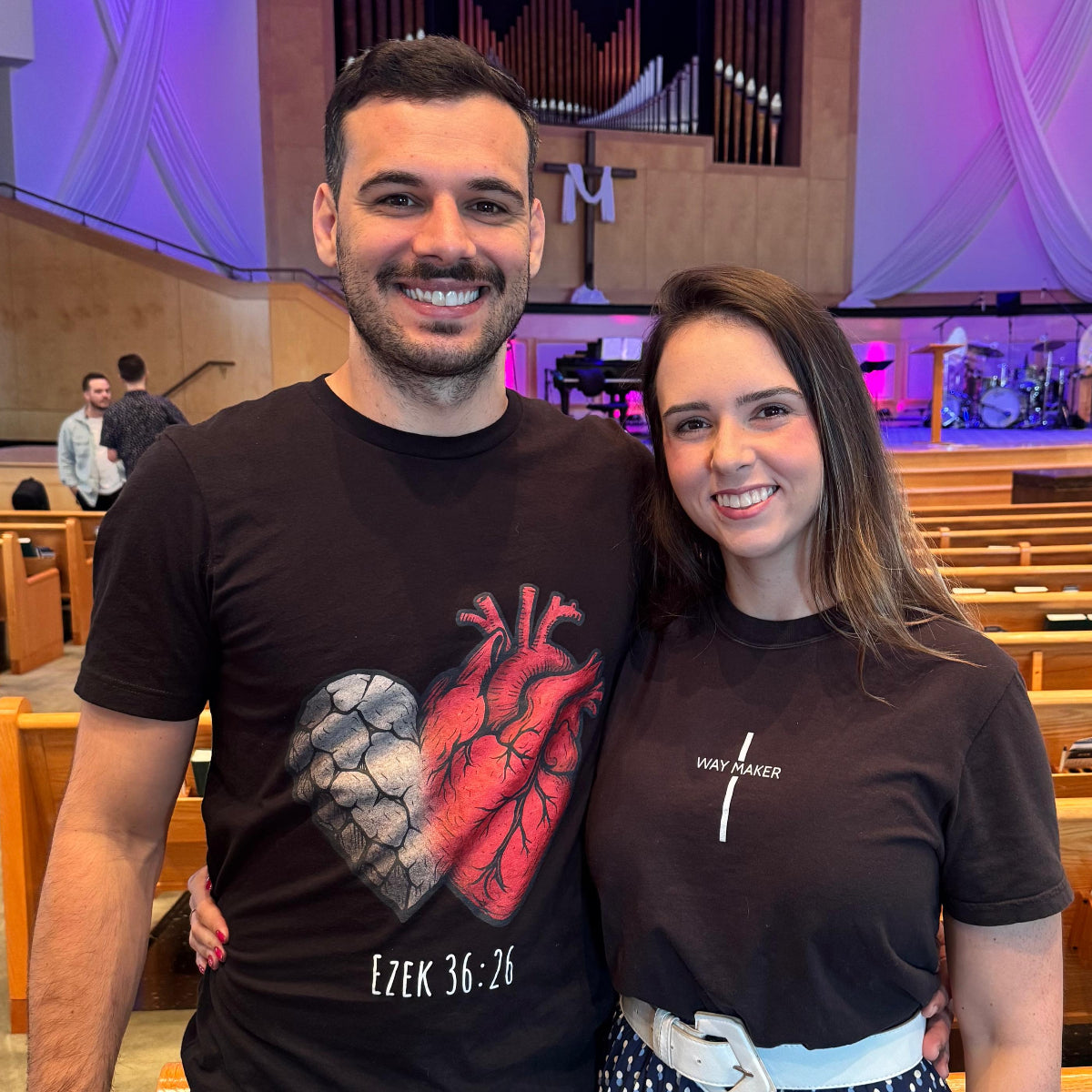 Two people standing in a church with wooden pews and a cross in the background.