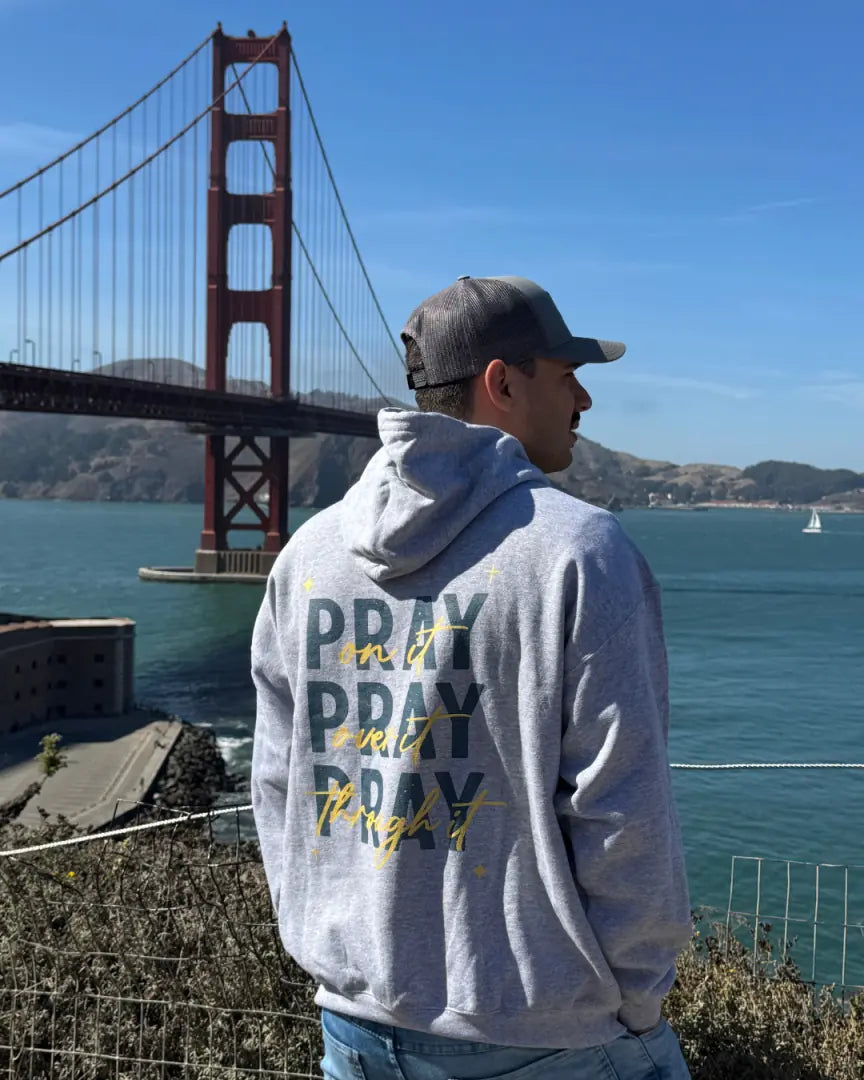 Person wearing a hoodie with 'PRAY' text in front of the Golden Gate Bridge.